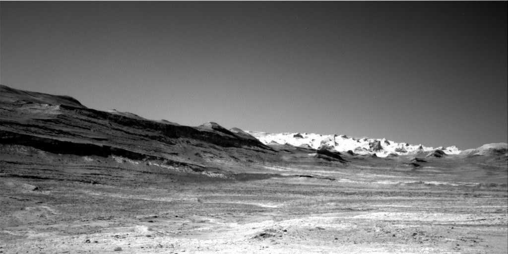 A grayscale landscape photo from the surface of Mars shows a broad area of flat terrain, colored light to medium gray, extending ahead from the foreground, leading to a slope that runs from the center-left of the frame downward toward middle of the image, where the ridge line runs more horizontally toward the center-right edge of the frame. Occasional outcrops along the ridge look like jagged teeth pointing up, and the sloped area at left looks like its made of numerous stacked layers of rock. In the distance beyond the ridge on the right side of the image a bright white series of peaks stands out, resembling a line of snow-capped mountains.